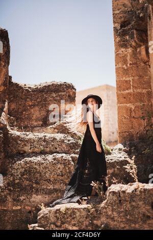 Vista laterale dell'elegante donna in un lungo vestito nero e cappello in piedi da un edificio in rovina e guardando indietro Foto Stock
