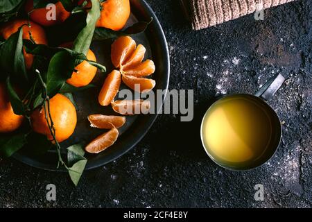 Vista dall'alto della tazza con succo di frutta fresca e piatto con i tangerini maturi posti sul tavolo nero ruvido vicino al tovagliolo Foto Stock