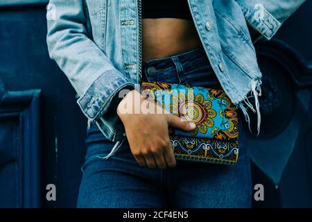 Crop African American Woman in jeans e giacca in denim in piedi con la frizione colorata alla moda in mano Foto Stock