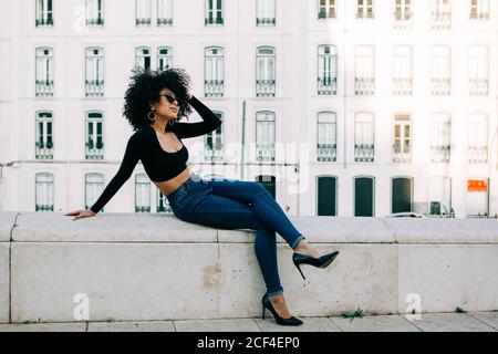 Giovane donna afroamericana alla moda in jeans e crop top seduto su parapetto di pietra e guardando sopra gli occhiali da sole Foto Stock