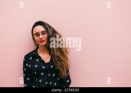 Donna adolescente affascinante in occhiali e camicia blu sorridente felicemente alla macchina fotografica in piedi contro la parete rosa Foto Stock