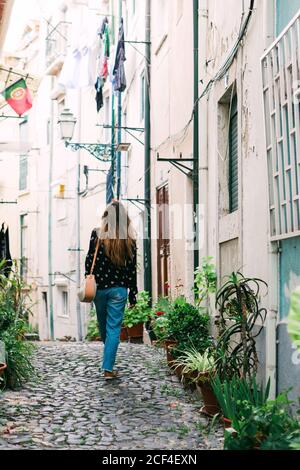 Vista sul retro di Casual Woman con borsa a piedi sulla strada lastricata stretta della città vecchia con vasi da fiori sui lati Foto Stock