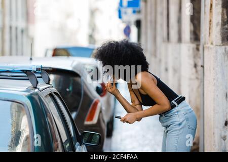 Vista laterale della giovane donna etnica guardando la finestra dell'auto e facendo trucco mentre si è in piedi su sfondo urbano Foto Stock