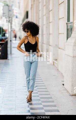 Giovane donna etnica in jeans e canotta a piedi e sorridente all'aperto Foto Stock