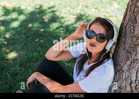 Donna pacifica in occhiali da sole e cuffie ascoltare musica mentre si siede sull'erba nel parco Foto Stock