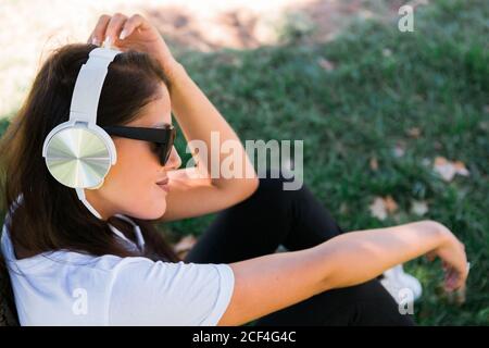 Donna pacifica in occhiali da sole e cuffie ascoltare musica mentre si siede sull'erba nel parco Foto Stock