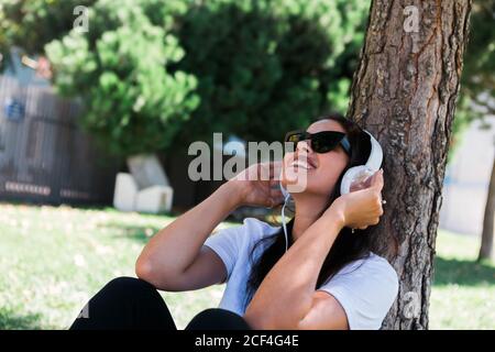 Donna pacifica in occhiali da sole e cuffie ascoltare musica mentre si siede sull'erba nel parco Foto Stock