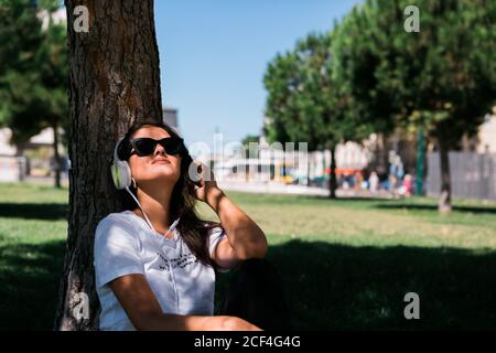 Donna pacifica in occhiali da sole e cuffie ascoltare musica mentre si siede sull'erba nel parco Foto Stock