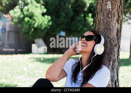 Donna pacifica in occhiali da sole e cuffie ascoltare musica mentre si siede sull'erba nel parco Foto Stock