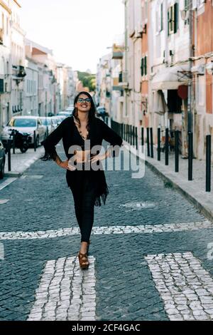 Giovane donna alla moda in vestito nero in piedi sul crosswalk con mani sui fianchi a Lisbona e sorridente Foto Stock