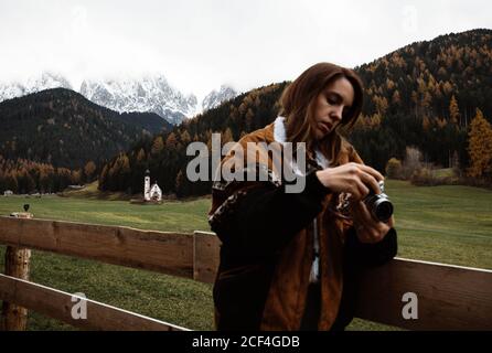 Giovane donna appoggiata ad una recinzione di legno usando una macchina fotografica vicino ad una piccola chiesa bianca rurale sul pendio della scogliera con il cielo della foresta autunnale e le montagne delle Dolomiti sullo sfondo Foto Stock