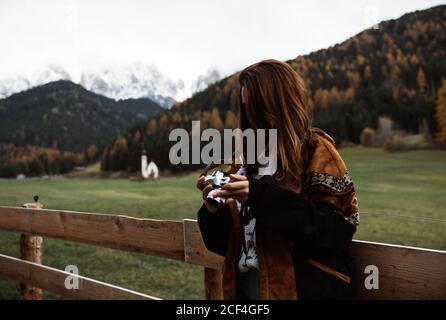 Giovane donna appoggiata ad una recinzione di legno usando una macchina fotografica vicino ad una piccola chiesa bianca rurale sul pendio della scogliera con il cielo della foresta autunnale e le montagne delle Dolomiti sullo sfondo Foto Stock