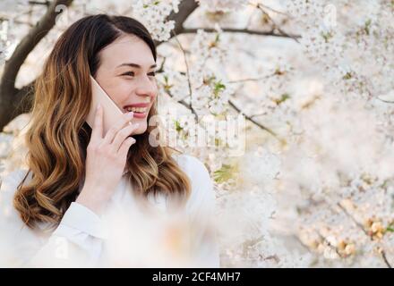 Giovane donna sorridente e parlando al telefono vicino ad alberi con fiori bianchi nella giornata di sole nel parco Foto Stock