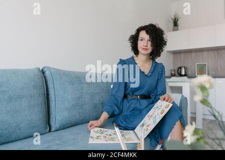 Bella donna giovane e bella bruna seduta sul divano e guardando tenere un album con i disegni a casa Foto Stock