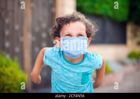 Ragazzo arrabbiato con maschera medica che piange perché non vuole indossare maschera medica. Concetto di bambini, salute ed emozioni Foto Stock