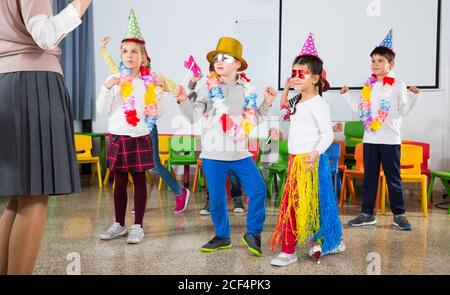 Contenti i bambini e insegnanti di sesso femminile in buffi cappelli e accessori di festa ballando insieme in auletta scuola Foto Stock