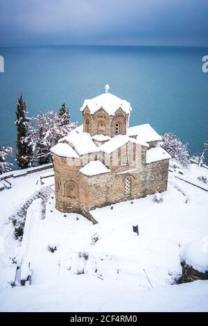 Antica chiesa ortodossa coperta di neve situata sulla costa situata Sulla riva del tranquillo lago Ohrid il giorno d'inverno in Macedonia del Nord Foto Stock