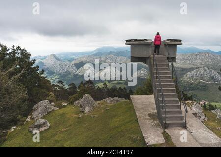 Vista posteriore di donna anonima in abiti casual in piedi osserva la piattaforma sul bordo della montagna e guarda favolosi Vista delle catene montuose in un giorno nuvoloso ad Alicante in Spagna Foto Stock