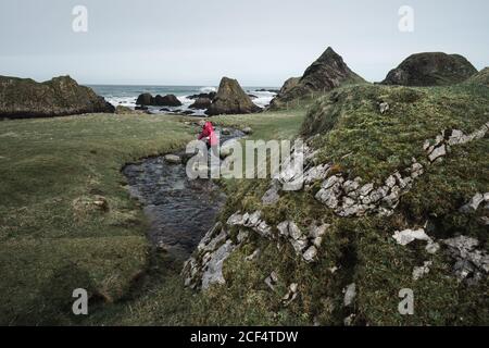 Dall'alto anonimo Woman godendo di un incredibile paesaggio panoramico dell'Irlanda del Nord durante il viaggio, mentre camminando vicino a un fiume poco profondo che scorre verso le acque della costa rocciosa Foto Stock
