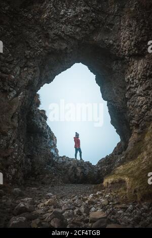 Vista laterale di femmina in abiti caldi in piedi sulla scogliera Bordo all'interno della grotta nel porto dell'Irlanda del Nord guardando lontano in mare Foto Stock