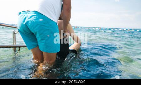 Uomo disabile in spiaggia che nuota su una sedia a rotelle con assistenza su una rampa accessibile. Foto Stock