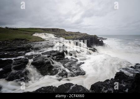 Onde di mare che si infrangono su rocce e si rompono a spruzzi In un giorno tempestoso con nuvole pesanti sulla costa dell'Irlanda del Nord Foto Stock