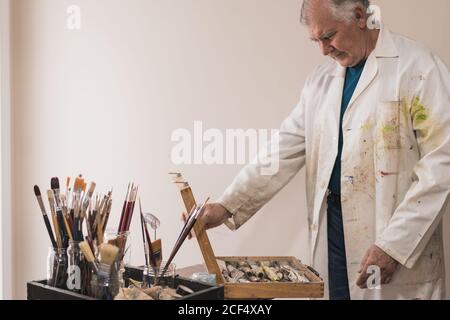 Artista maschile invecchiato in cappotto da lavoro colorato in piedi vicino al tavolo con set di pennelli e scatola di apertura con vernici mentre scegliere le vernici e lavorare in studio moderno Foto Stock