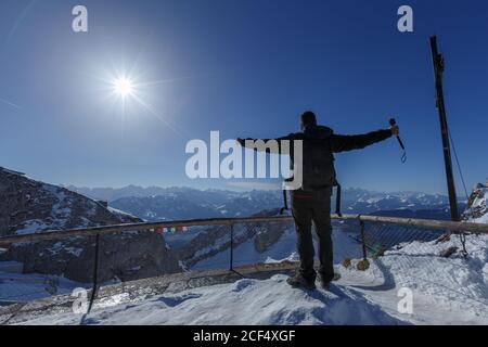 Vista posteriore dell'uomo con zaino che tiene le braccia in piedi sulla terrazza della montagna innevata con un panorama incredibile sotto la luce del sole Foto Stock