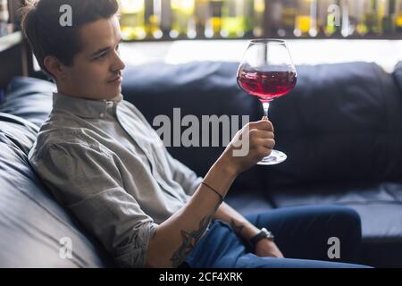 Focalizzato giovane maschio sommelier in suite guardando il vino rosso in vetro su sfondo bianco Foto Stock