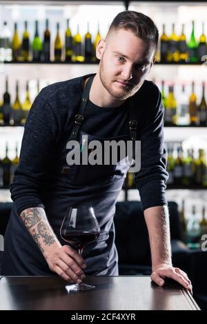 Ritratto di un sommelier in piedi in un negozio che tiene un bicchiere di vino e mostra il pollice in su, durante una degustazione Foto Stock