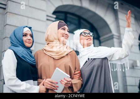 Tre sorelle musulmane che camminano. La prima donna con occhiali punta al cielo. Due ragazze che guardano, all'aperto. Vicino all'edificio Foto Stock