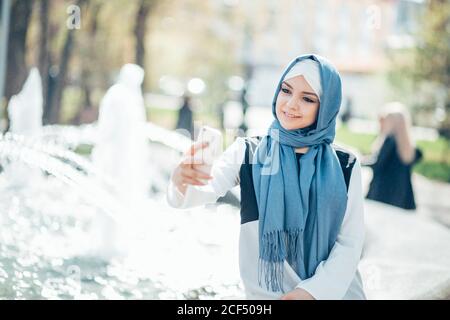 Ritratto bella donna in hijab facendo selfie da smartphone all'aperto. Sullo sfondo della fontana Foto Stock