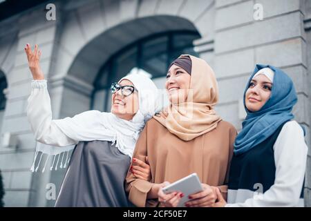 Tre sorelle musulmane che camminano. La prima donna con occhiali punta al cielo. Due ragazze che guardano, all'aperto. Vicino all'edificio Foto Stock