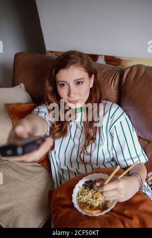 Da sopra giovane redhead Donna mangiare ramen e cambiare canali in TV mentre si siede sul divano durante il pranzo a casa Foto Stock