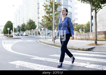 Imprenditore femminile positivo in abito elegante tenendo la mano in tasca mentre camminate sul crosswalk sulla strada della città moderna Foto Stock