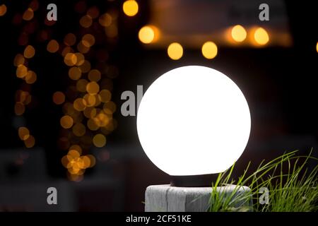 la luce rotonda bianca della strada risplende sulla colonna con l'erba verde, sullo sfondo caldo bokeh circoli diffusi da ghirlande di notte su sfondo scuro, vacanza p Foto Stock