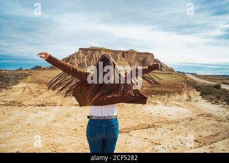 Vista posteriore di anonimo viaggiatore donna in abbigliamento casual in piedi con le mani sollevate vicino a scogliera marrone e cielo blu sullo sfondo a Bardenas Reales, Navarra, Spagna Foto Stock