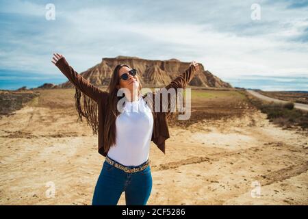 Soddisfatto viaggiatore femminile in abbigliamento casual e occhiali da sole in piedi con le mani sollevate e sorridente con scogliera marrone e cielo blu sullo sfondo a Bardenas Reales, Navarre, Spagna Foto Stock