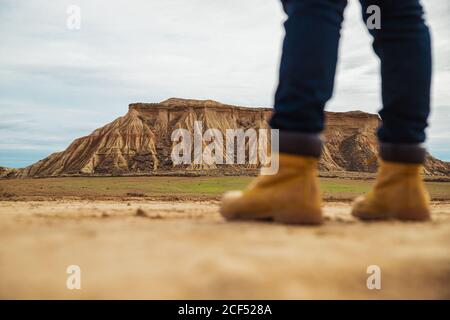 Gambe sul viaggiatore senza volto in stivali marroni e jeans blu in piedi su strada di sabbia sporca con montagna e cielo su sfondo sfocato a Bardenas Reales, Navarre, Spagna Foto Stock
