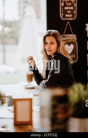 Vista laterale di capelli lunghi alla moda bella bionda Donna bere da un bicchiere di delizioso caffè schiumoso mentre si guarda la macchina fotografica Foto Stock