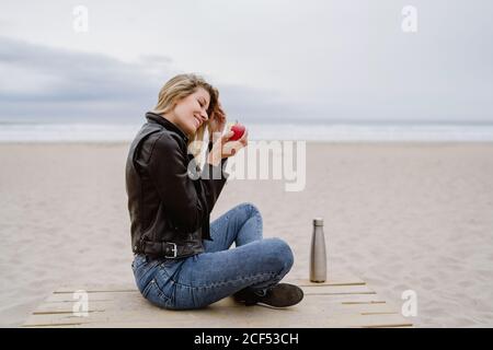 Vista laterale della donna bionda alla moda con berretto nero e giacca in pelle che mangia mela rossa matura sulla spiaggia sabbiosa Foto Stock