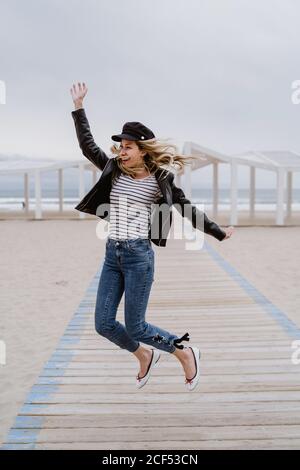 Donna bionda trendy in giacca nera saltando allegro con le braccia sollevate su molo di legno bianco con oceano sullo sfondo Foto Stock