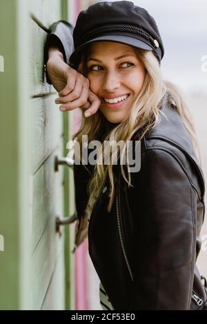 Bionda alla moda dai capelli lunghi Donna in berretto nero e giacca in pelle sorridente e poggiante sulle pareti delle cabine in legno della spiaggia Foto Stock