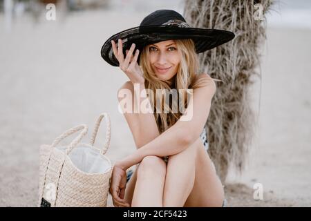 Bella donna bionda in cappello nero seduta sulla sabbia con borsa estiva e guardando la macchina fotografica Foto Stock