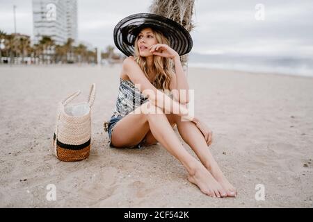 Bella donna bionda in cappello nero seduta sulla sabbia con borsa estiva e guardando lontano pensivo Foto Stock