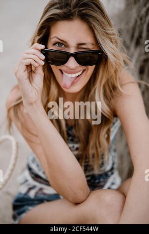 Primo piano ritratto di bella donna bionda in occhiali da sole neri seduti sulla sabbia e guardando la macchina fotografica che si stacca dalla lingua e che si inocchiava Foto Stock