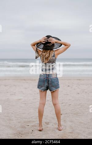 Vista posteriore di attraente donna slim indossare top e shorts in piedi sulla spiaggia sabbiosa con cappello nero Foto Stock