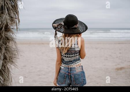 Vista posteriore di attraente donna slim indossare top e shorts in piedi sulla spiaggia sabbiosa con cappello nero Foto Stock