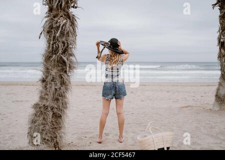 Vista posteriore di attraente donna slim indossare top e shorts in piedi sulla spiaggia sabbiosa con cappello nero Foto Stock