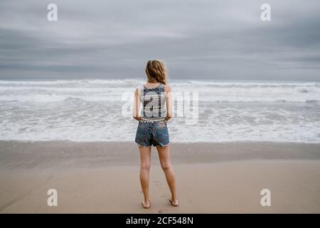 Vista posteriore di anonimo attraente slim Woman indossare top e shorts in piedi sulla spiaggia sabbiosa Foto Stock